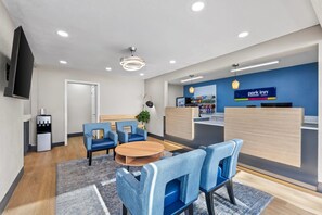 Zona de recepción de hotel con pared azul, mostrador de recepción, área de descanso con sillas, máquina de café y televisor montado en la pared.