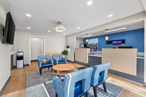 Zona de recepción de hotel con pared azul, mostrador de recepción, área de descanso con sillas, máquina de café y televisor montado en la pared.