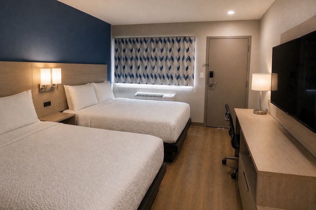 A hotel room with two beds, a TV, a desk with a lamp, a window with patterned curtains, and a door.