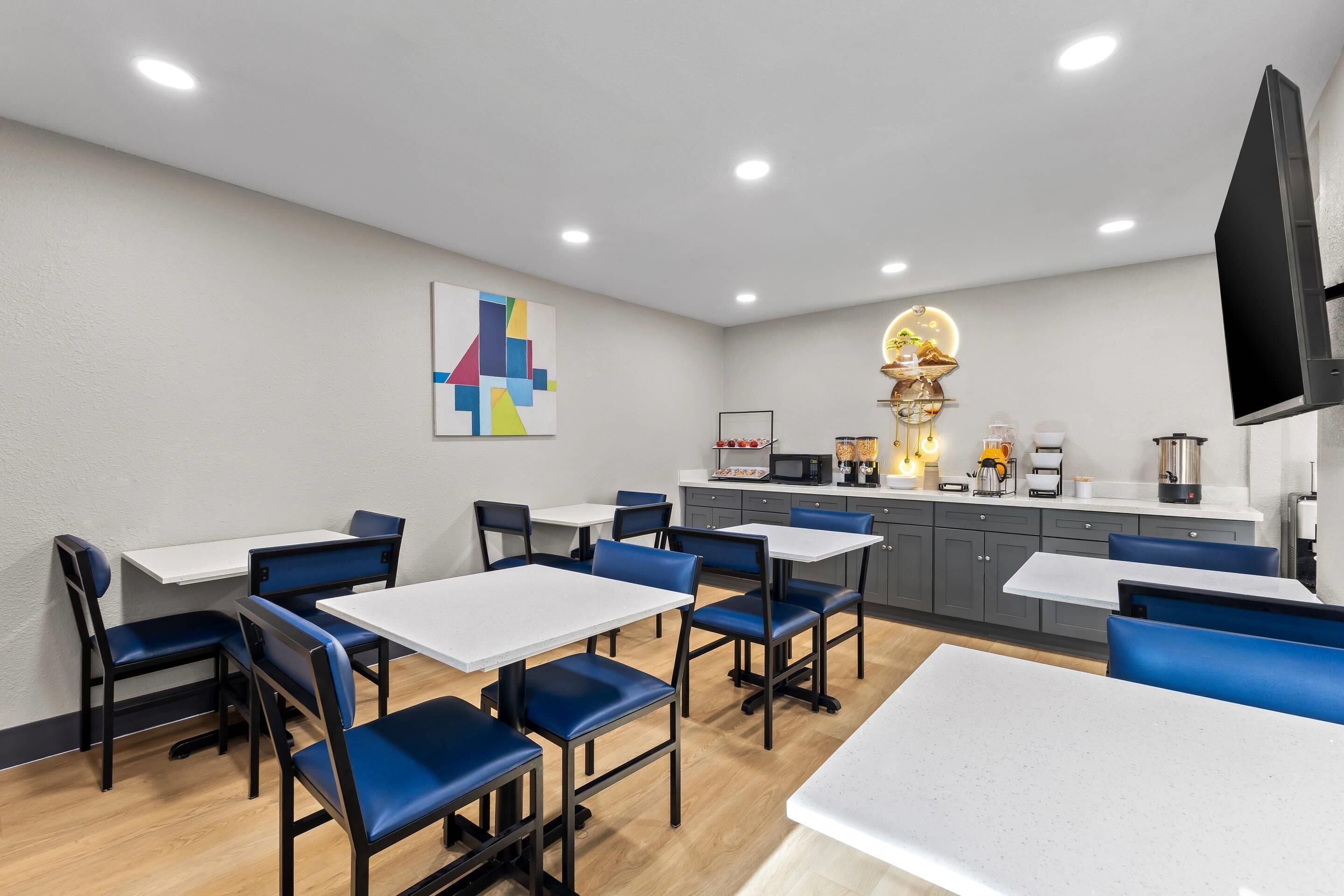 Een moderne eetruimte met witte tafels en blauwe stoelen, een keuken met apparatuur, en een grote flatscreen televisie aan de muur.