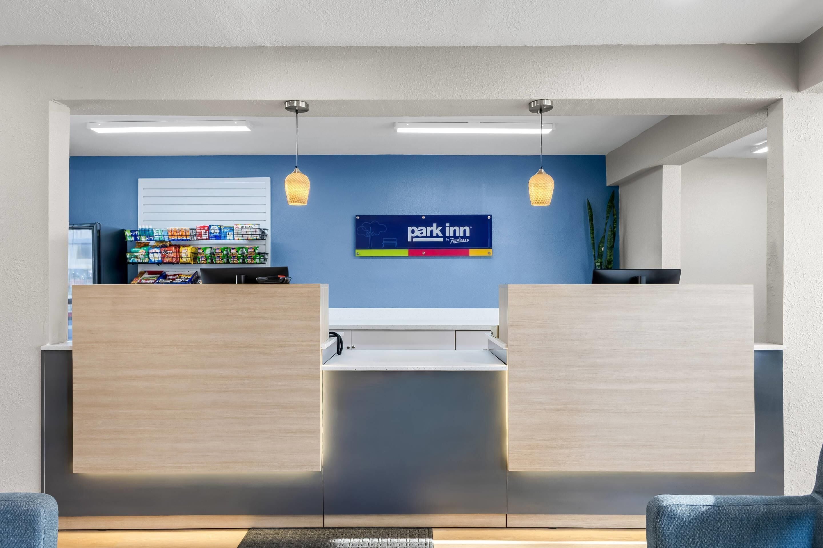 Uma área de recepção com parede azul, uma placa 'Park Inn' e um balcão de lanches com diversos produtos embalados.