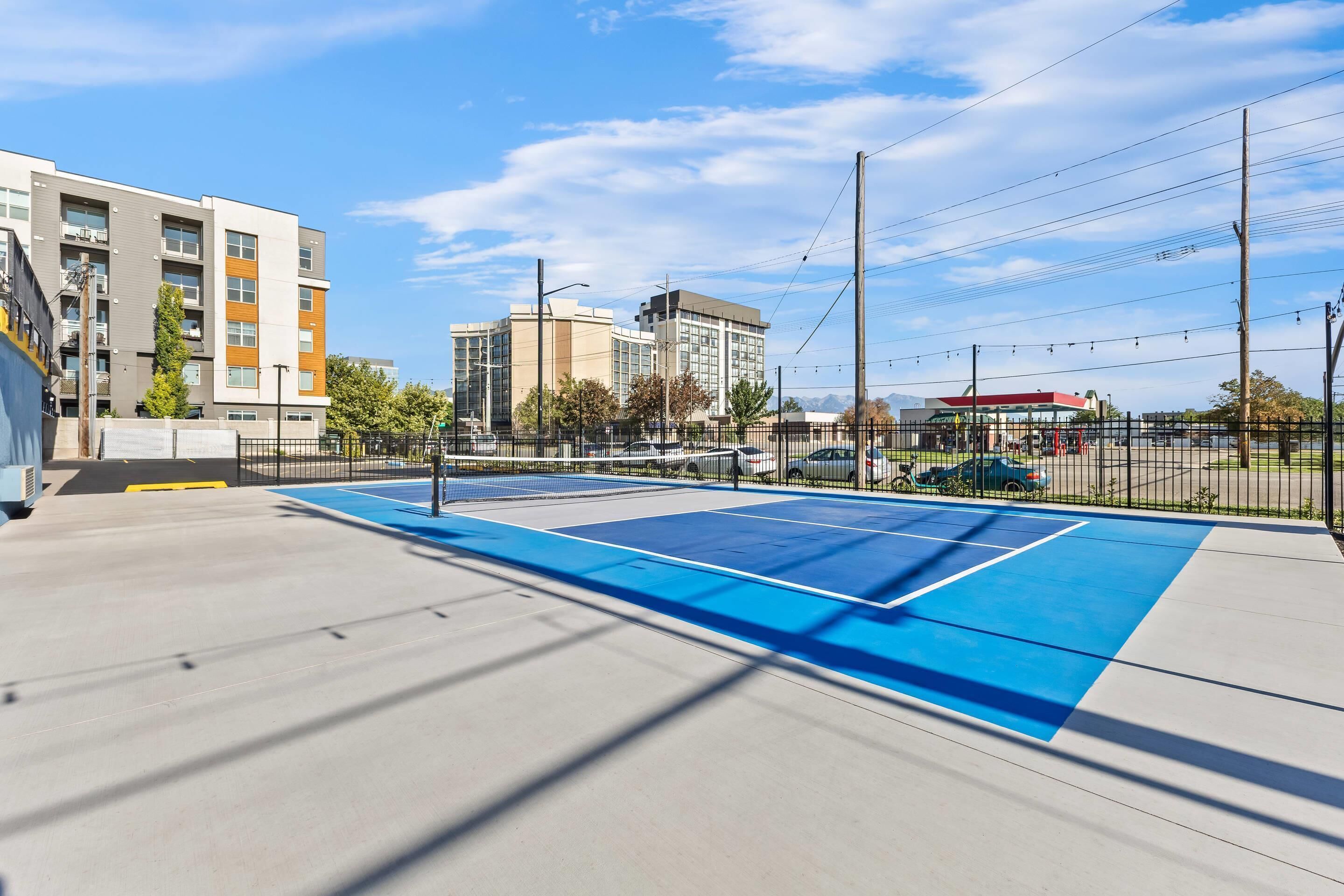 A blue tennis court with a fence, surrounded by buildings and a clear sky.