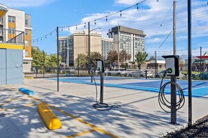 Zona de carga para vehículos eléctricos al aire libre, con cielo despejado, edificios modernos al fondo y un espacio de estacionamiento azul.