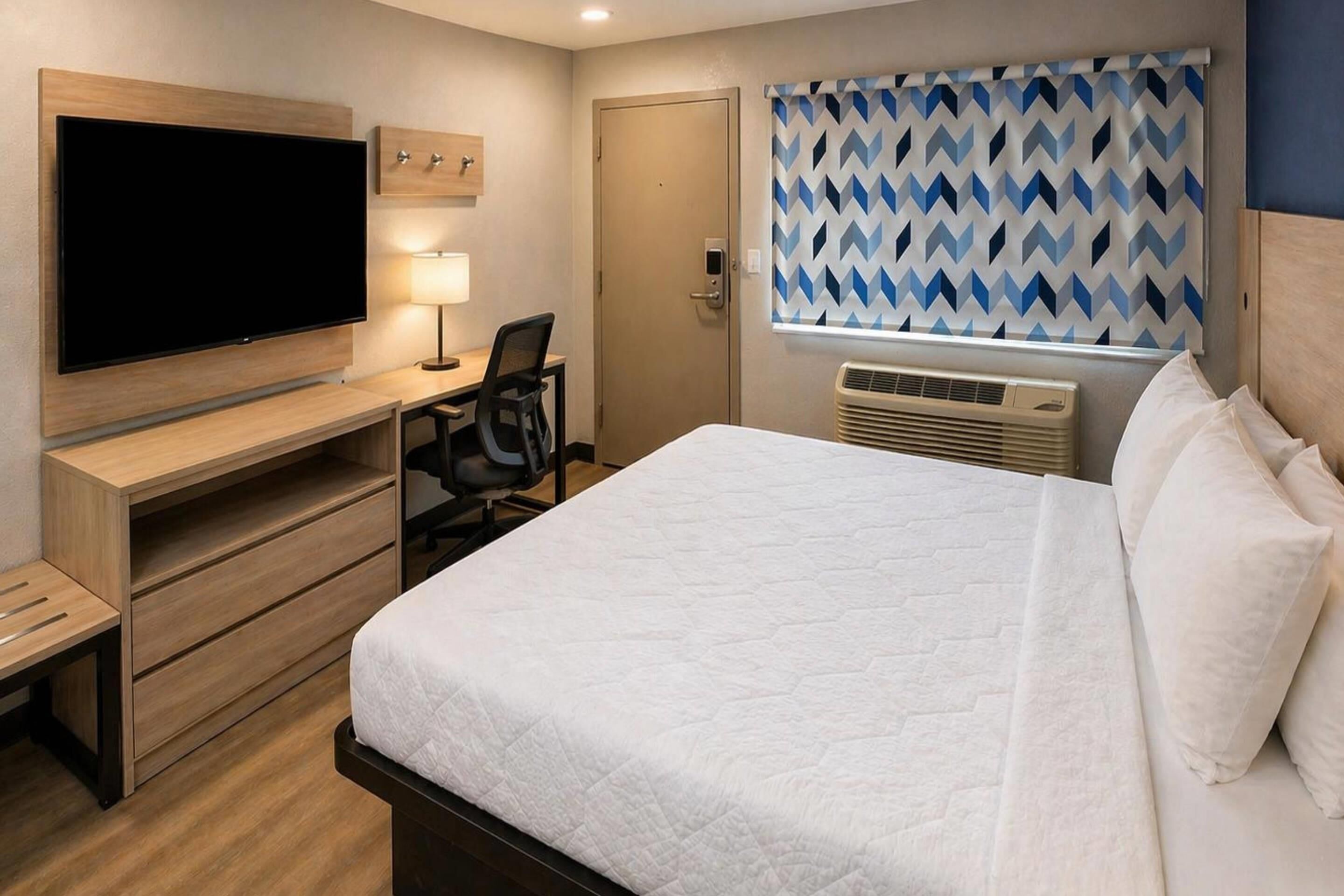 Habitación de hotel con cama, un escritorio con silla, televisor, ventana y cortinas estampadas, y una puerta.