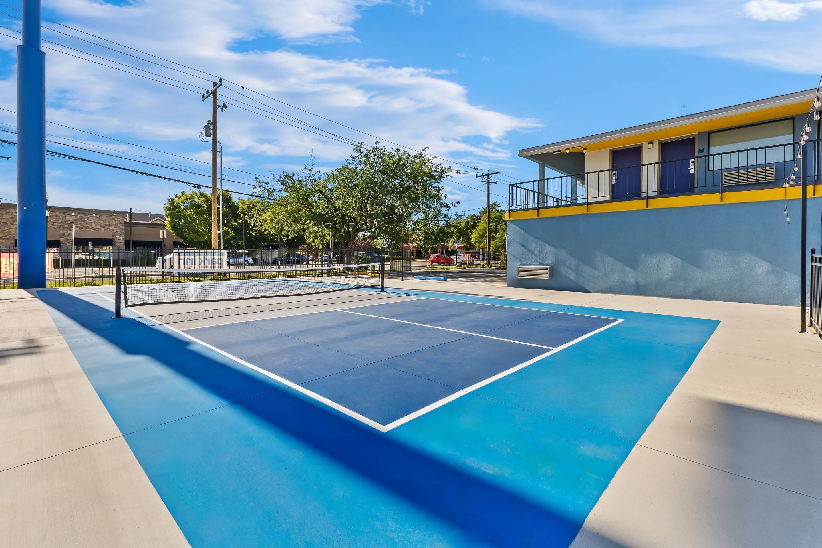 An outdoor tennis court with a blue surface, surrounded by a fence, and adjacent to a building with a yellow and blue facade.
