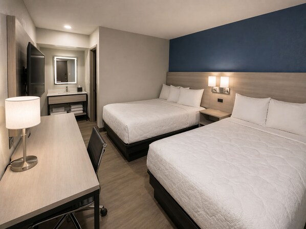 A hotel room with two beds, a desk, a mirror, and a bathroom visible in the background.