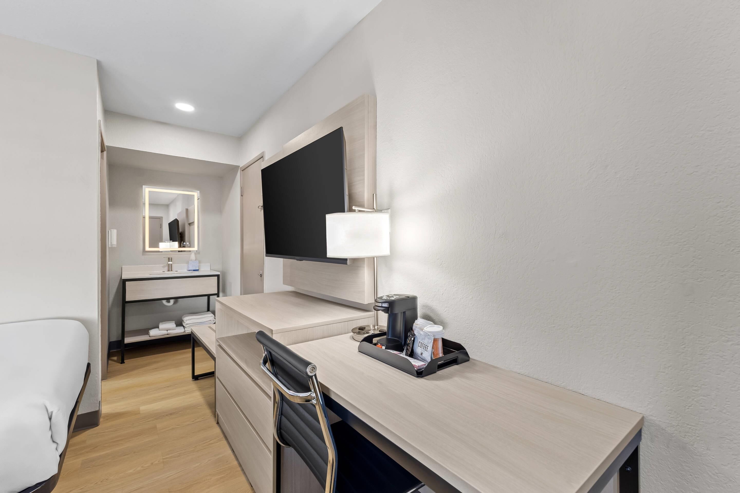 Quarto de hotel com secretária, cadeira, televisão, espelho e máquina de café.