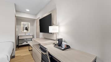 A hotel room with a desk, a chair, a TV, a mirror, and a coffee machine.