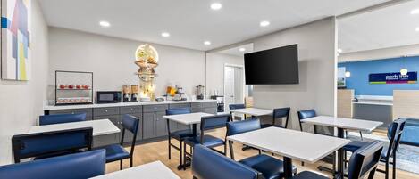 A modern dining area with tables and chairs, a kitchen area with appliances, and a flat-screen TV mounted on the wall.