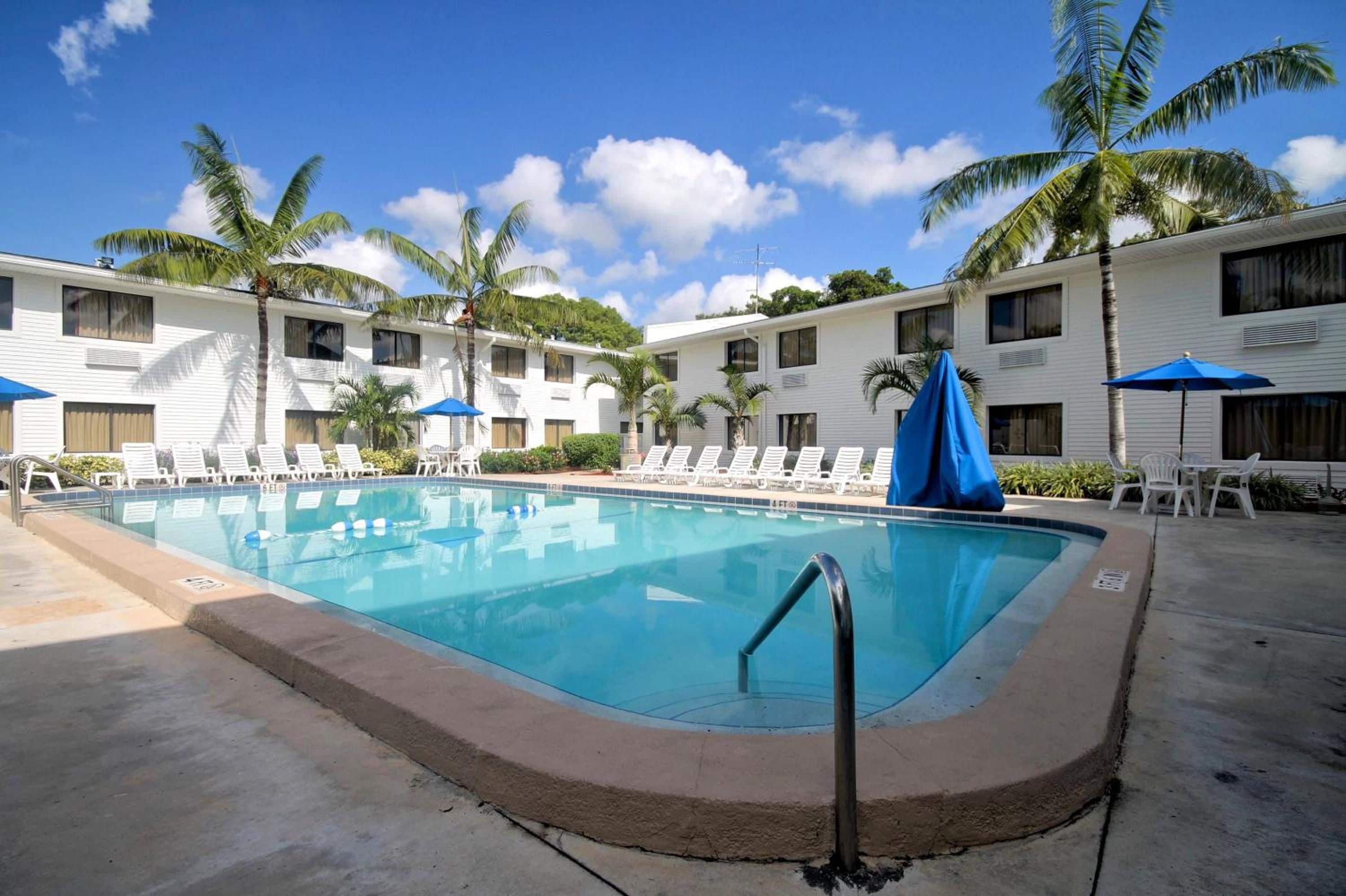 A swimming pool with lounge chairs and umbrellas, surrounded by palm trees and a multi-story building.