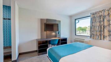 A hotel room with a bed, a desk with a chair, a TV, and a window with patterned curtains.