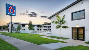 A motel with a sign indicating Wi-Fi availability, a modern building with a glass door, and a well-maintained lawn. - Motel 6 Fort Lauderdale, FL (Fort Lauderdale)