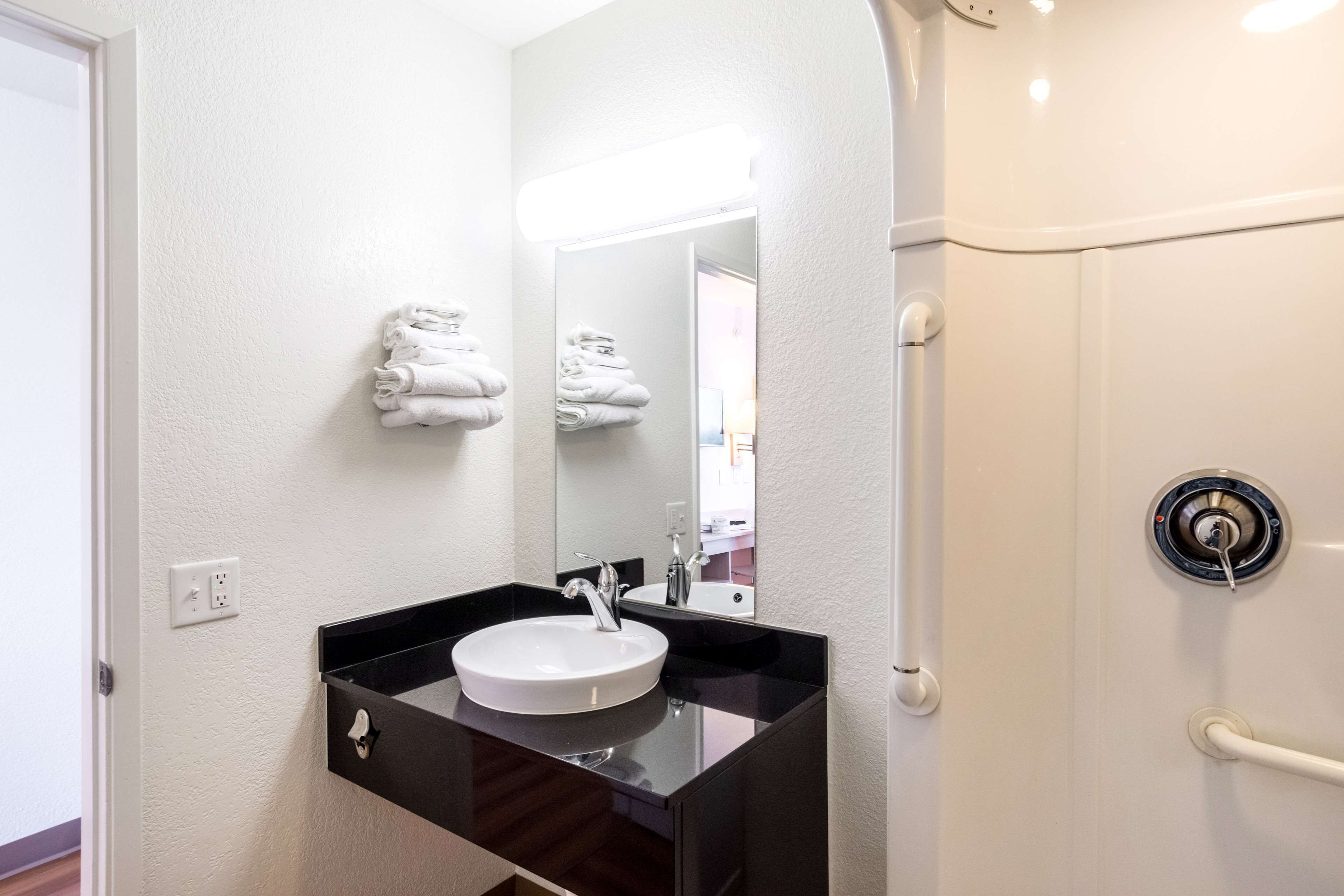 Een badkamer met een wit wastafel, een zwart aanrechtblad en een spiegel. Handdoeken liggen opgestapeld aan de muur en er is een douchedeur met een handgreep.