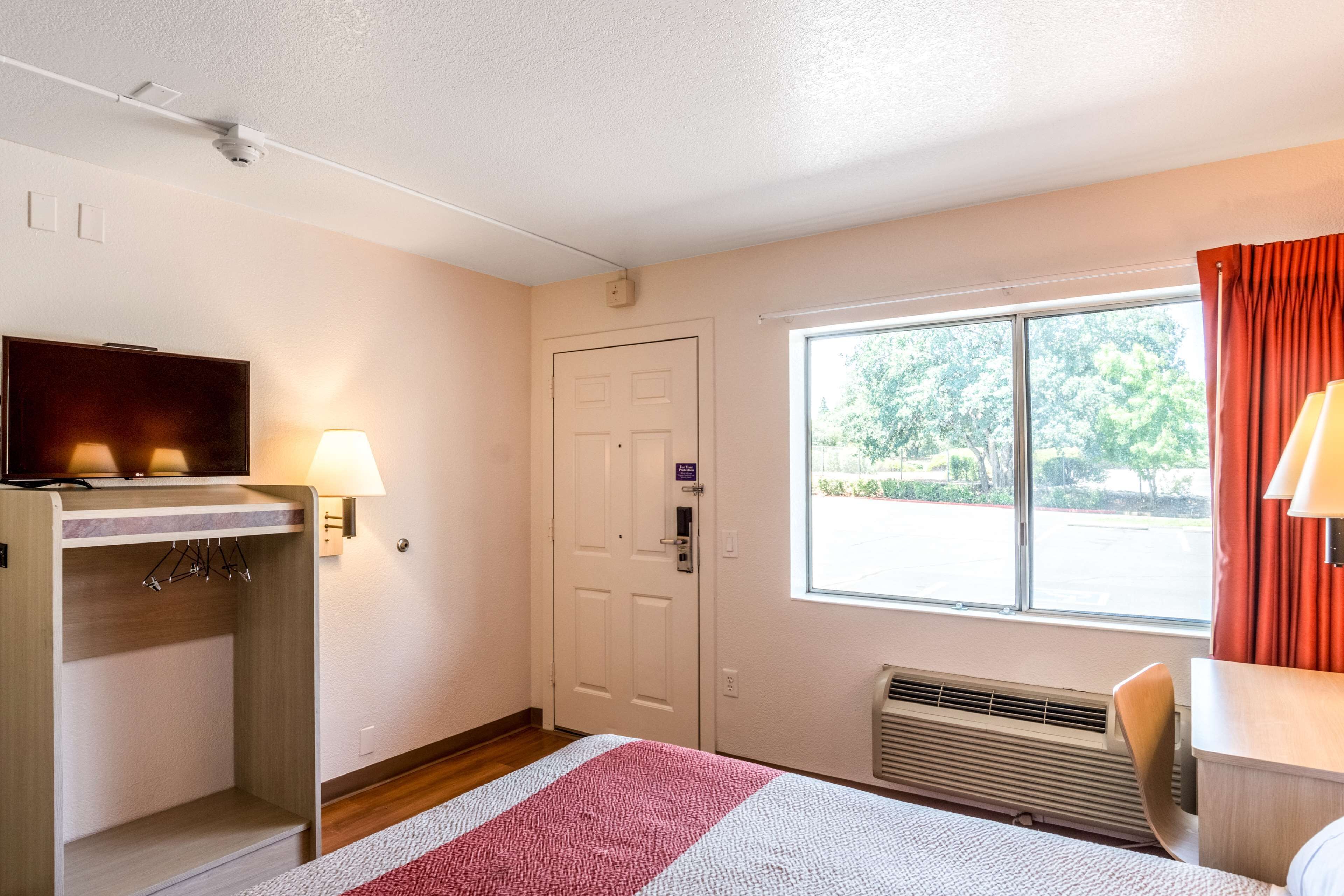 A hotel room with a bed, a desk with a lamp, a television, a door, and a window with a view of trees.