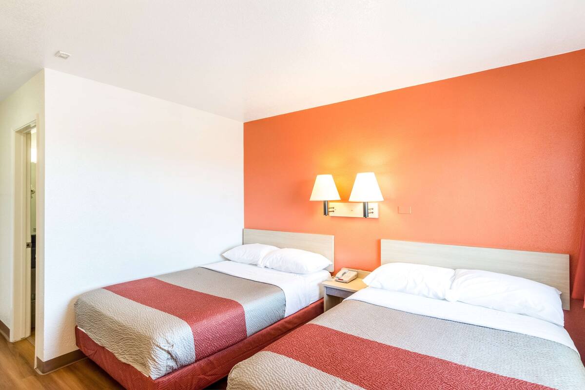 A hotel room with two beds, each with a red and grey bedspread, white pillows, and a nightstand with a lamp.