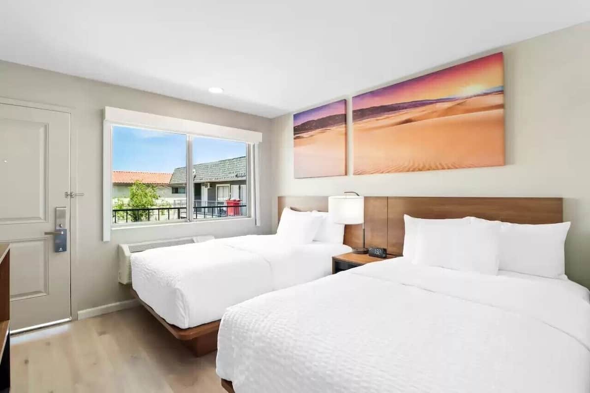 A hotel room with two beds, a window with a view of buildings, and a painting on the wall.