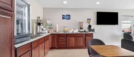 A modern kitchen with wooden cabinets, a refrigerator, a coffee machine, and a flat-screen TV mounted on the wall.
