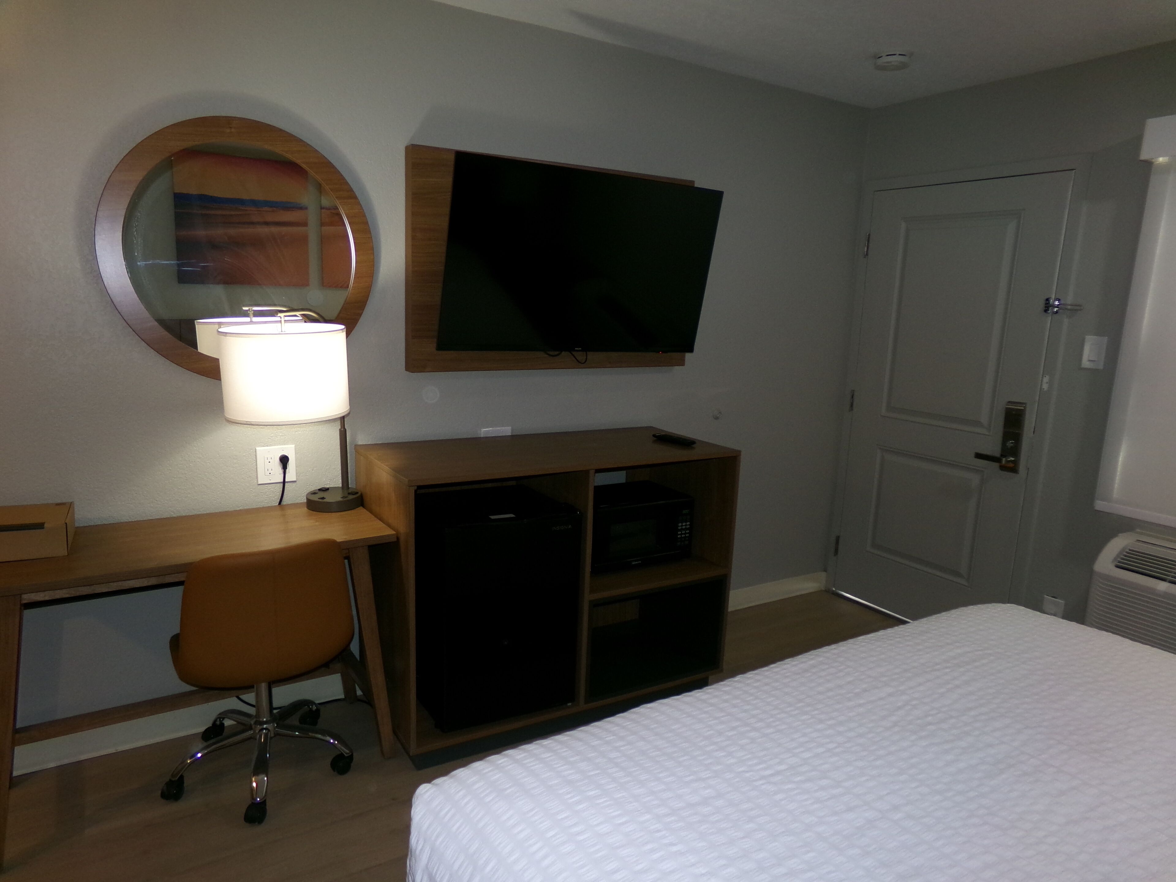 A hotel room with a bed, a desk with a chair, a television mounted on the wall, and a door.