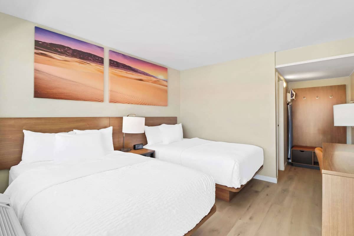 A hotel room with two beds, a wooden headboard, a nightstand with a lamp, and a large painting of a desert landscape.