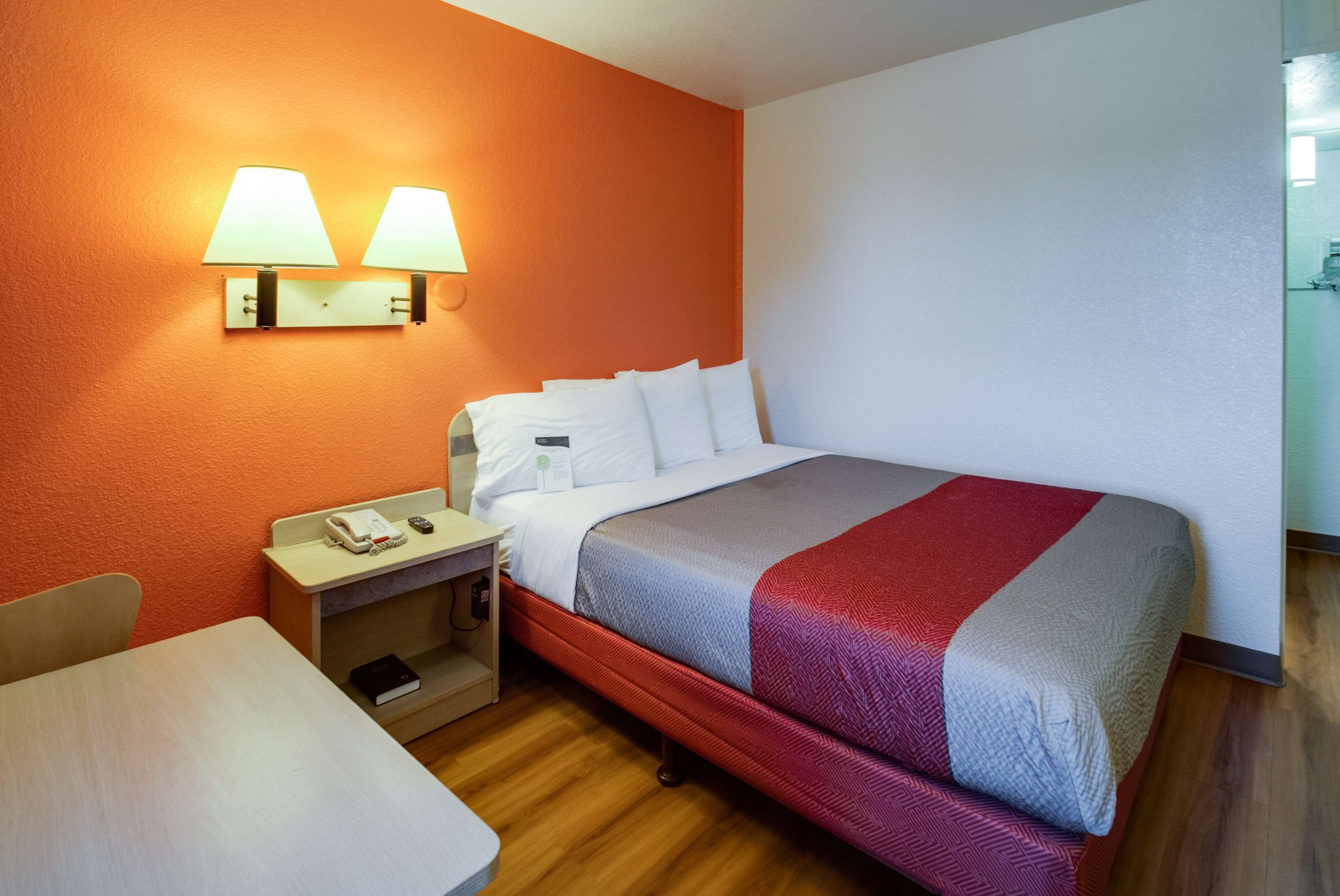 A hotel room with a bed, a nightstand with a lamp, a table, and a wall-mounted light fixture.