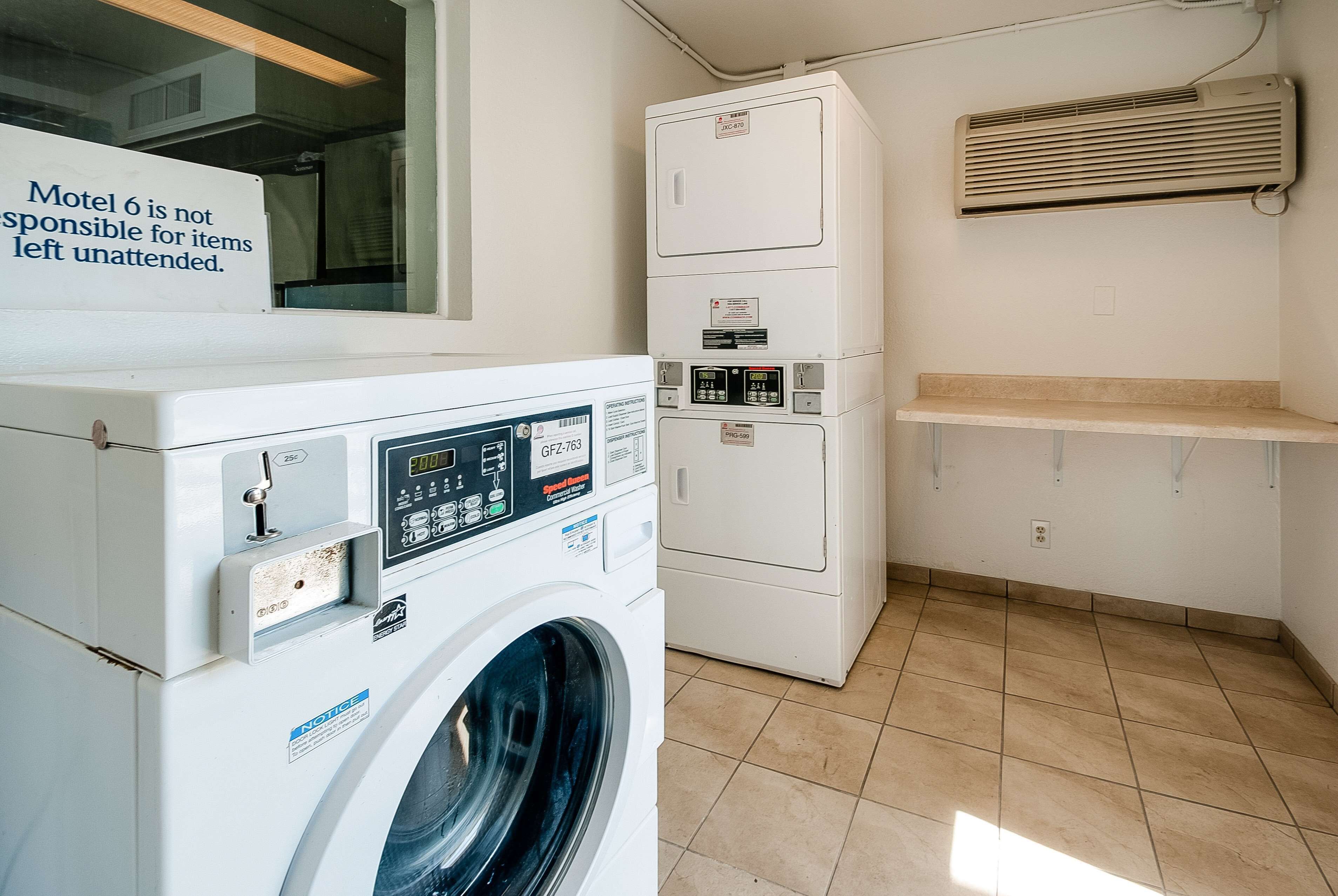 A laundry area with a front-loading washing machine, stacked dryers, and a sign stating 'Motel 6 is not responsible for items left unattended.'