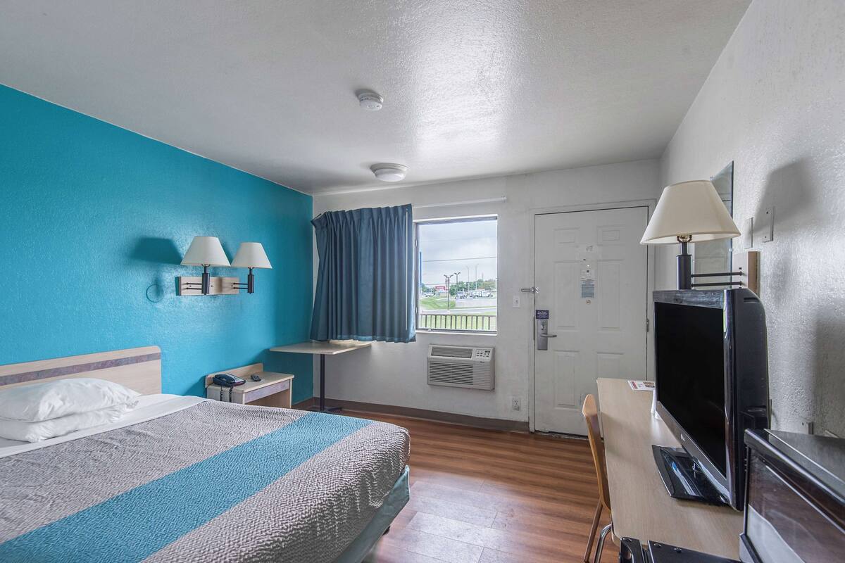 A hotel room with a bed, a desk with a TV, a window with curtains, and a door.