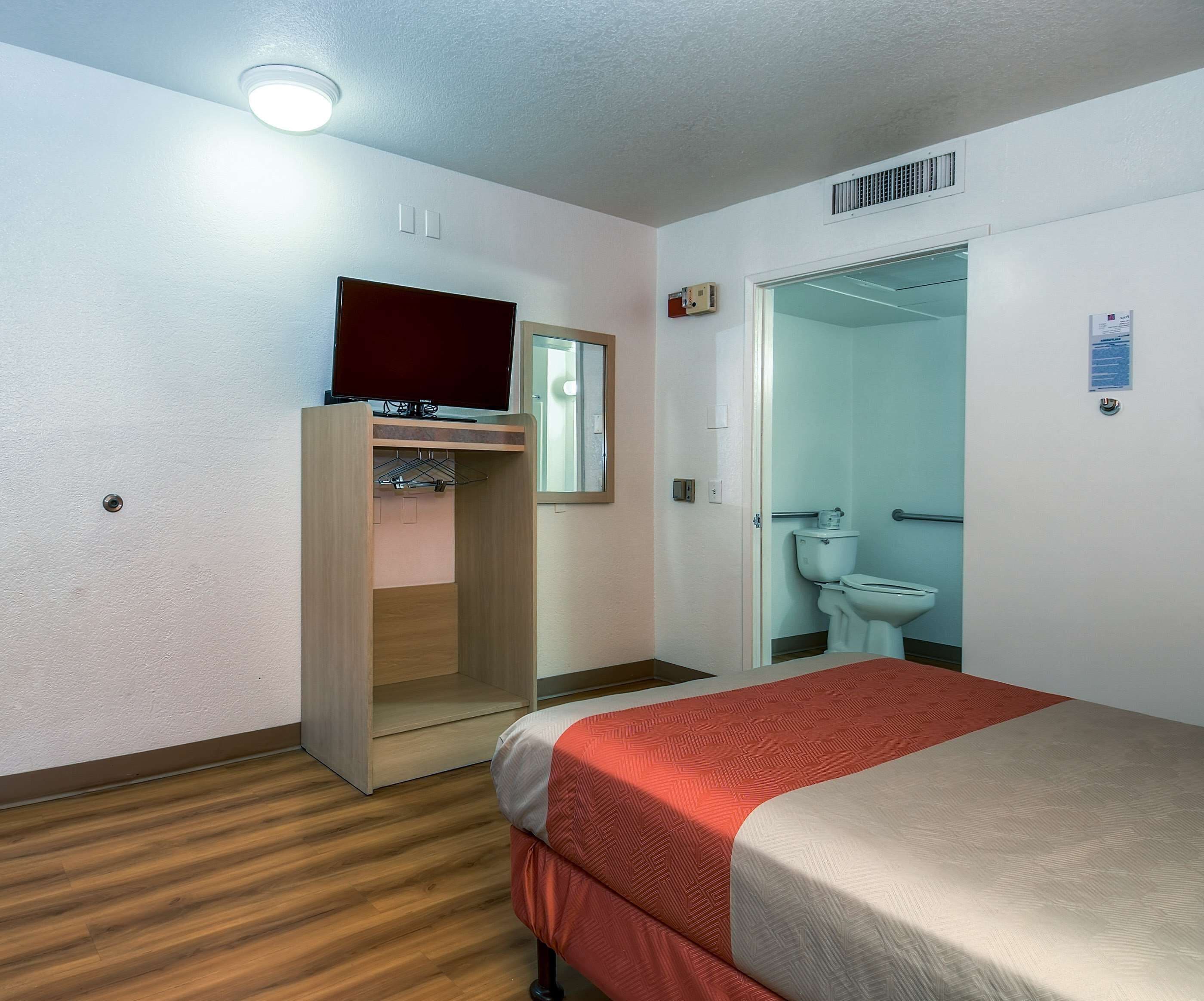 A hotel room with a bed, a television on a stand, a mirror, a wardrobe, and a bathroom with a toilet and sink.
