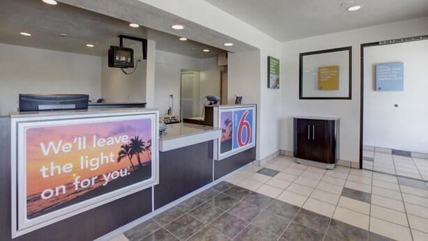 A reception area with a counter, a digital screen, and a sign that reads "We'll leave the light on for you." - Motel 6 Corona, CA (Corona)
