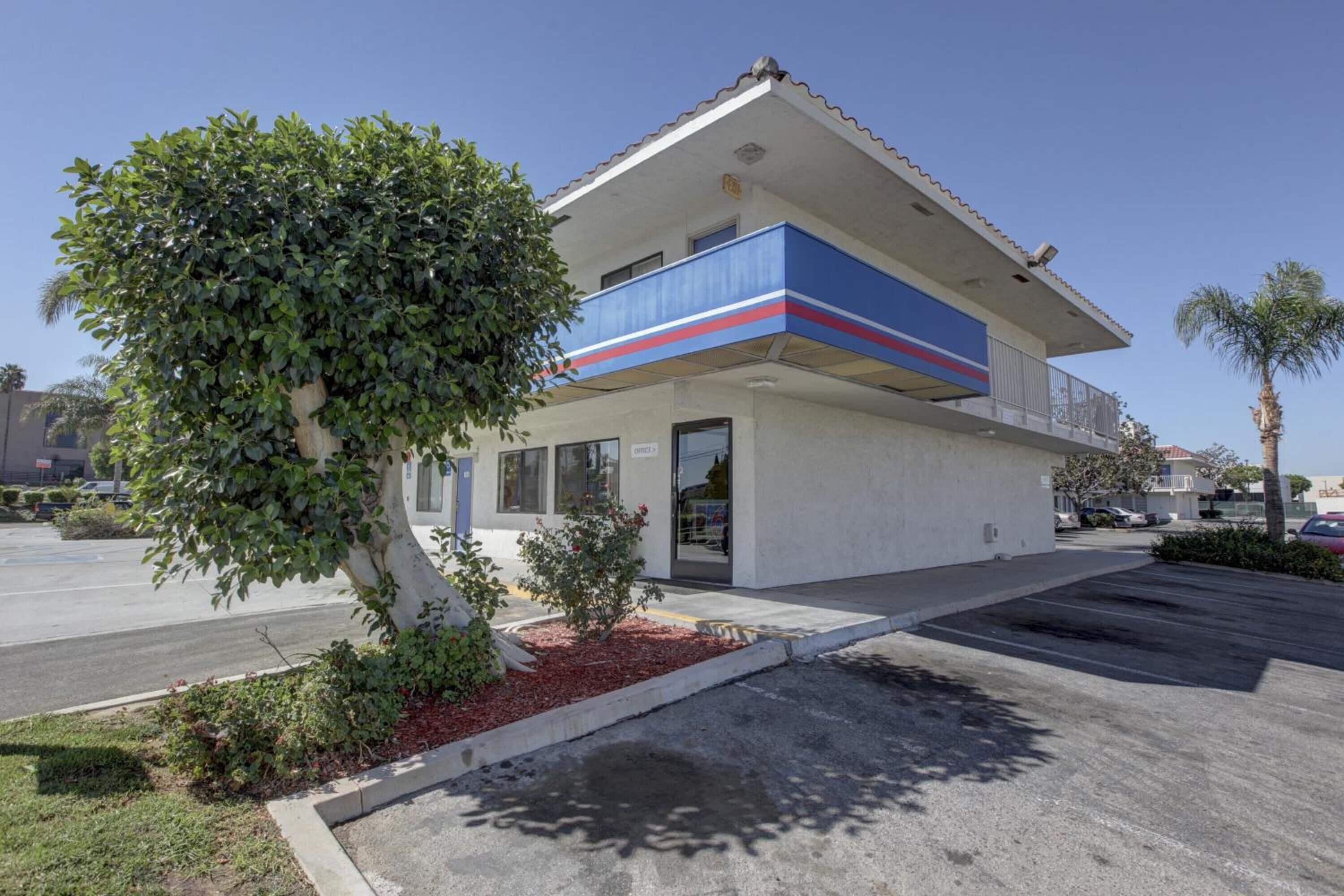 A two-story building with a blue and red stripe on the facade, a covered parking area, and a small tree in front.