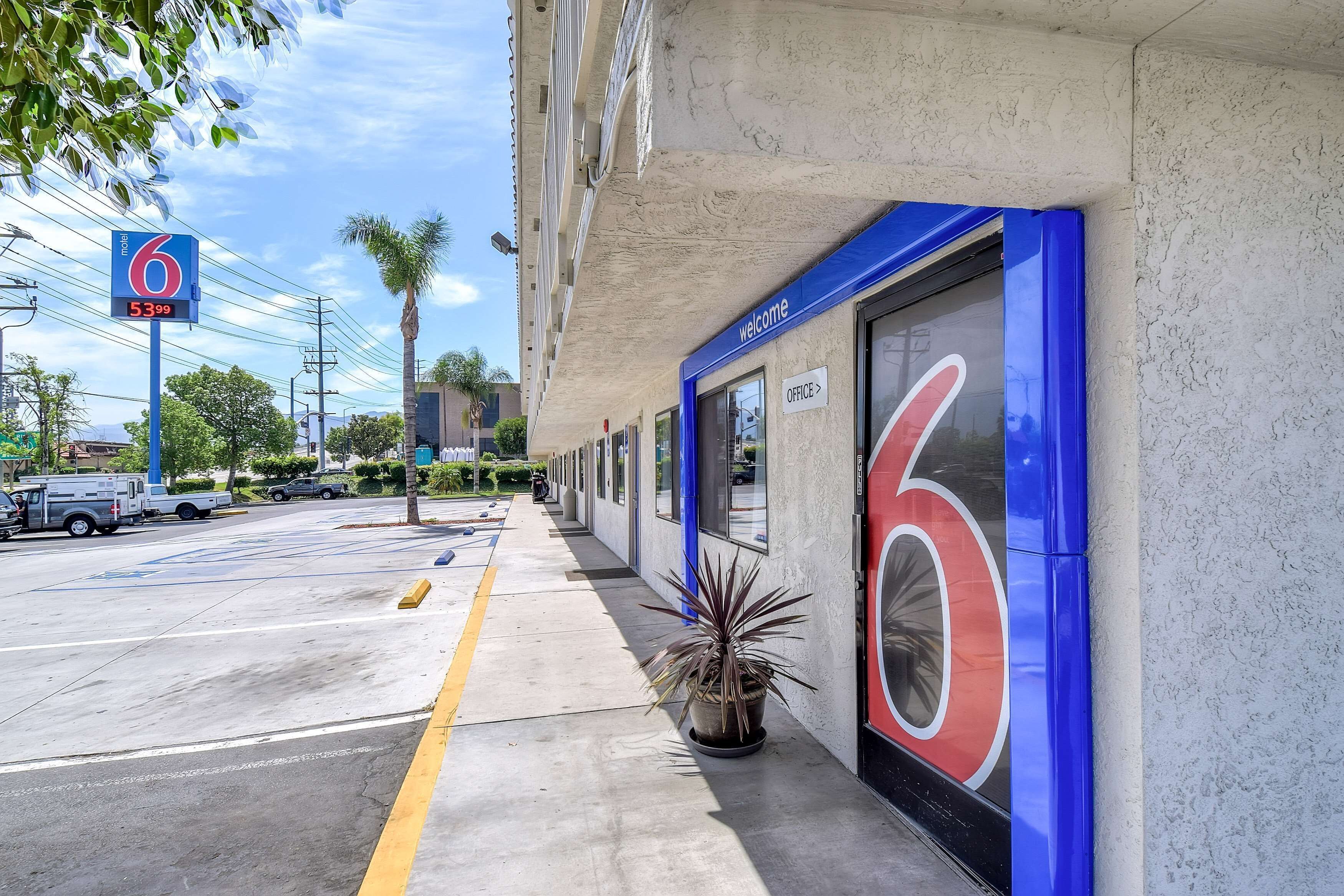Entrada de un hotel con un cartel grande con el número '6', un letrero de bienvenida y un logotipo del número '6' en el edificio.