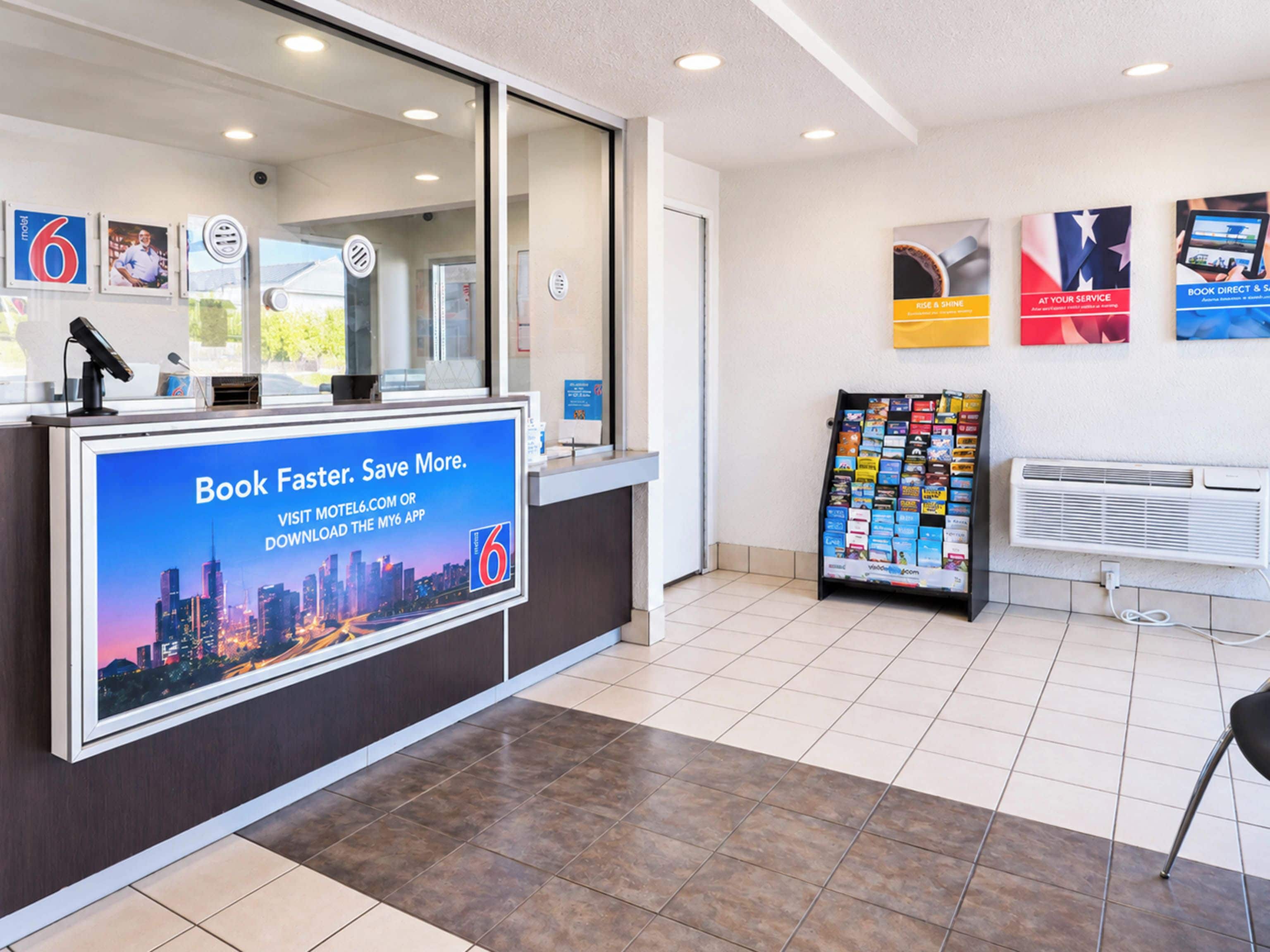 A hotel reception area with a digital screen displaying a promotional message, a bulletin board with various notices, and a tiled floor.