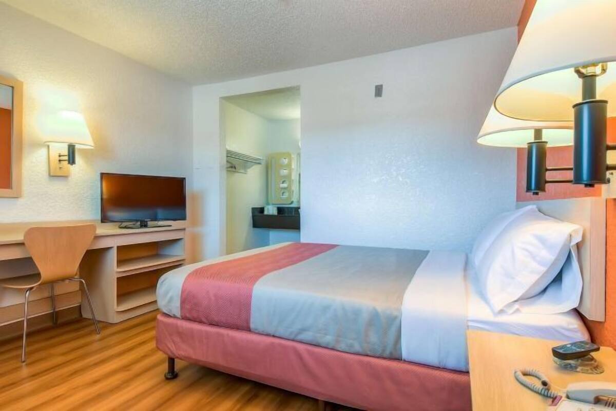 A hotel room with a bed, desk, chair, television, and a lamp.