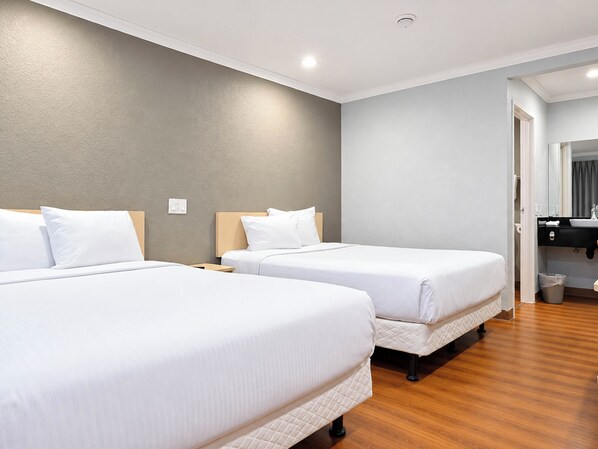 A hotel room with two beds, a wooden floor, a grey wall, and a bathroom with a mirror and sink. - Motel 6 Rowland Heights, CA - Los Angeles - Pomona (Rowland Heights)