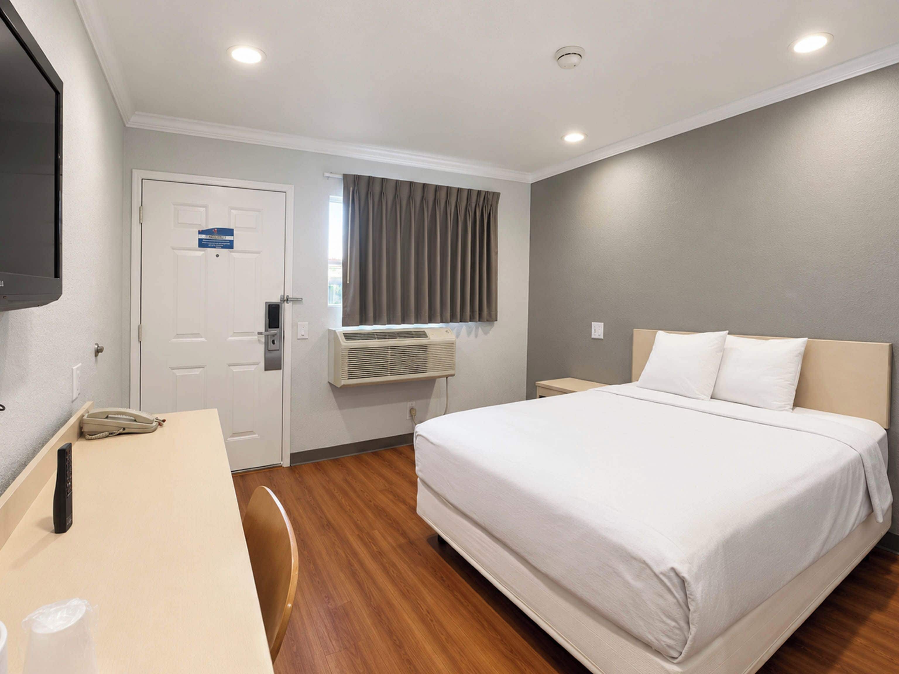 A hotel room with a bed, a desk, a chair, a television, and an air conditioning unit.