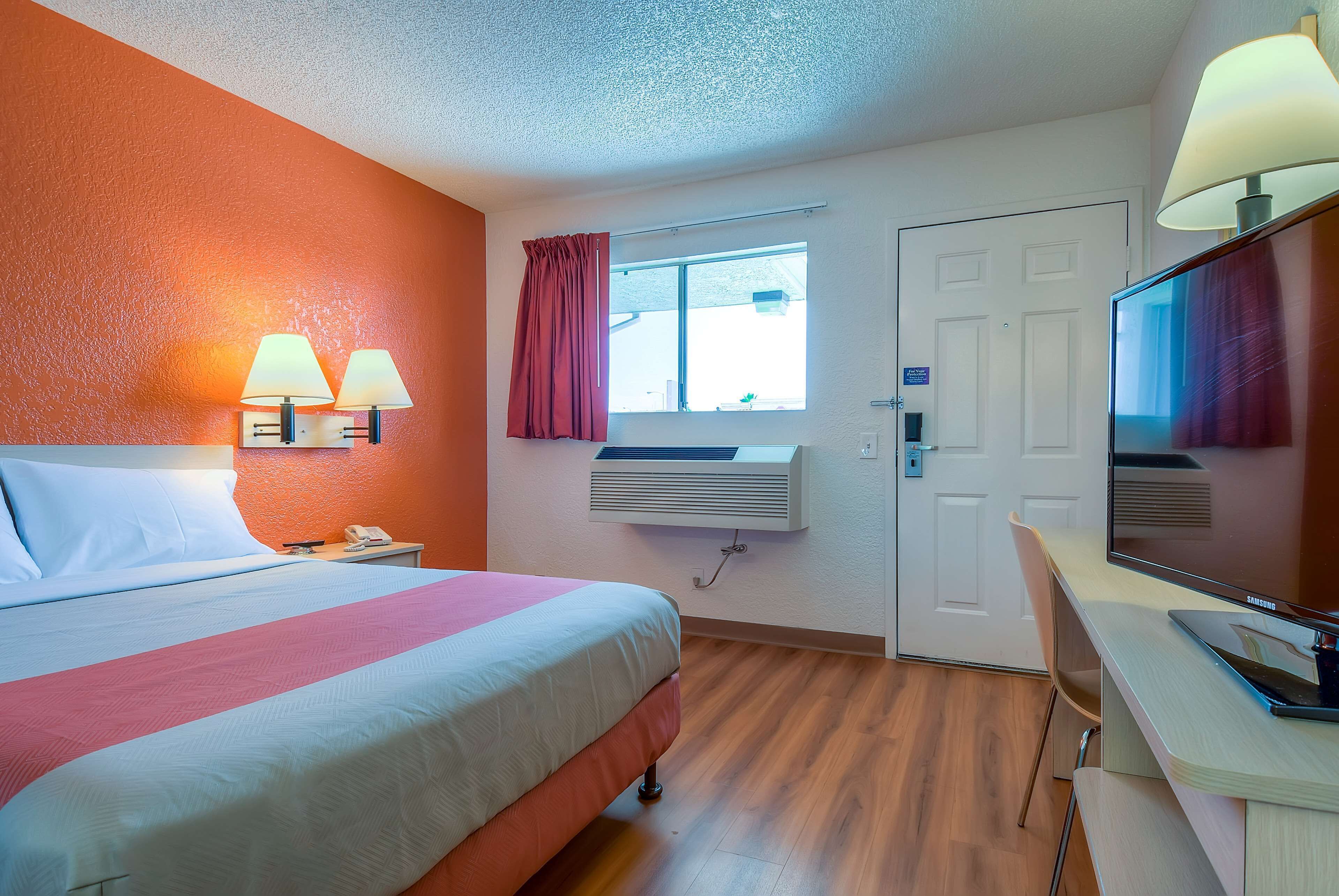 Habitación de hotel con cama, lámparas de noche, escritorio con computadora, televisor, ventana con cortinas y aire acondicionado.