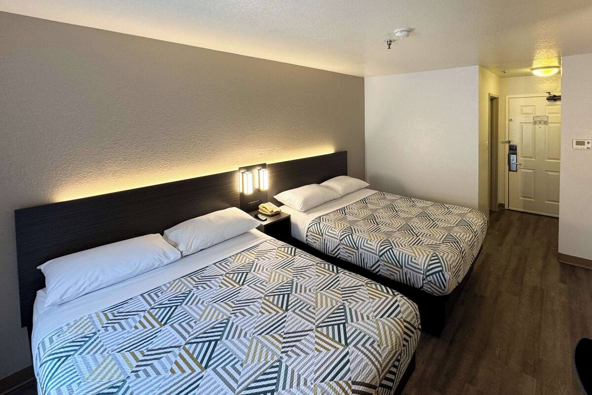 A hotel room with two beds, each with patterned bedding, a nightstand with a lamp, and a door leading to another room.