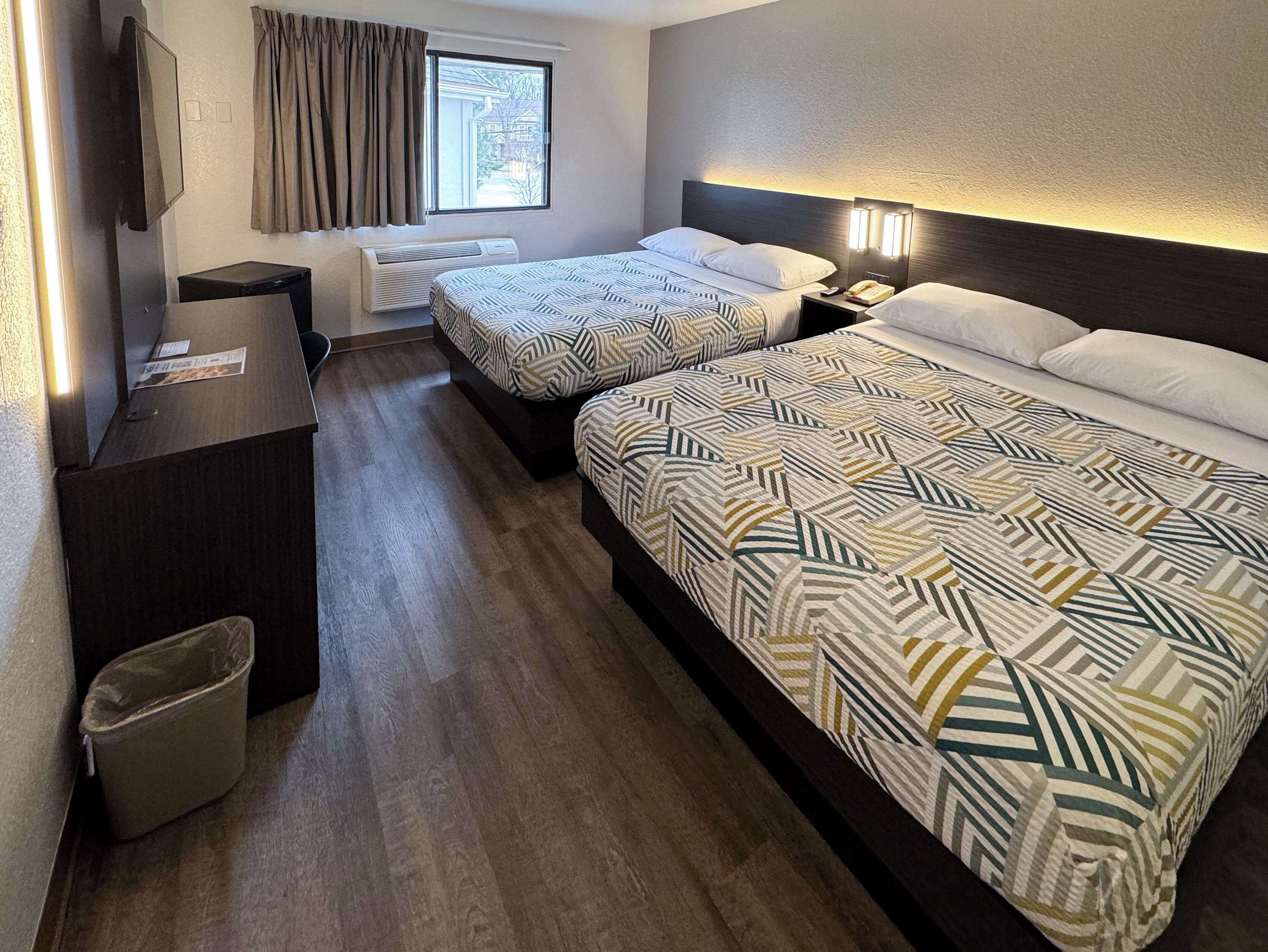 A hotel room with two beds, a desk, a trash bin, a window with curtains, and a wall-mounted TV.
