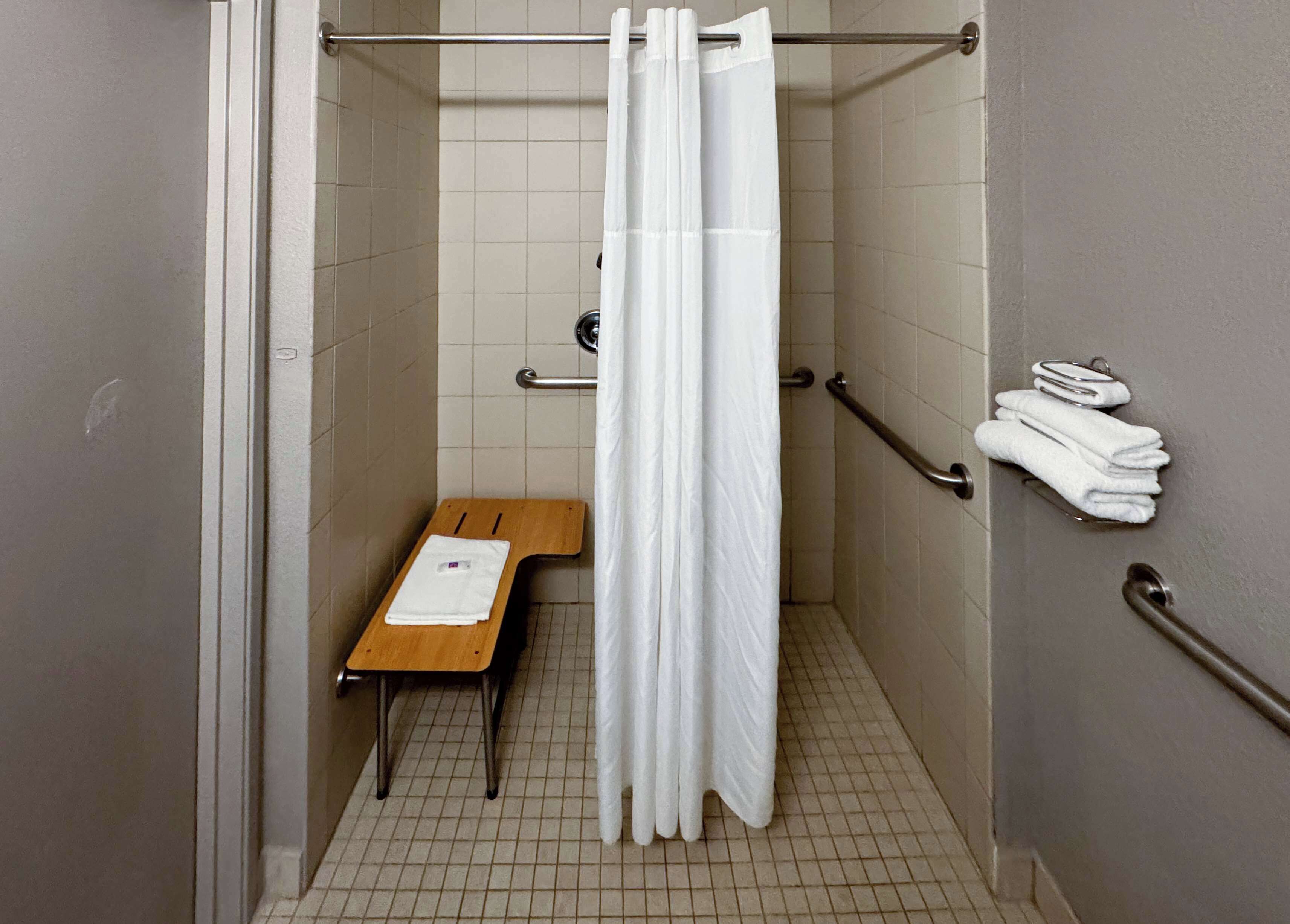 A small, tiled shower stall with a wooden bench, a white shower curtain, and a towel rack with folded towels.
