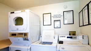A laundry area with multiple washing machines and dryers, including models 619 and 796. There are informational signs on the wall, one of which states 'Motel 6 is not responsible for items left unattended.'