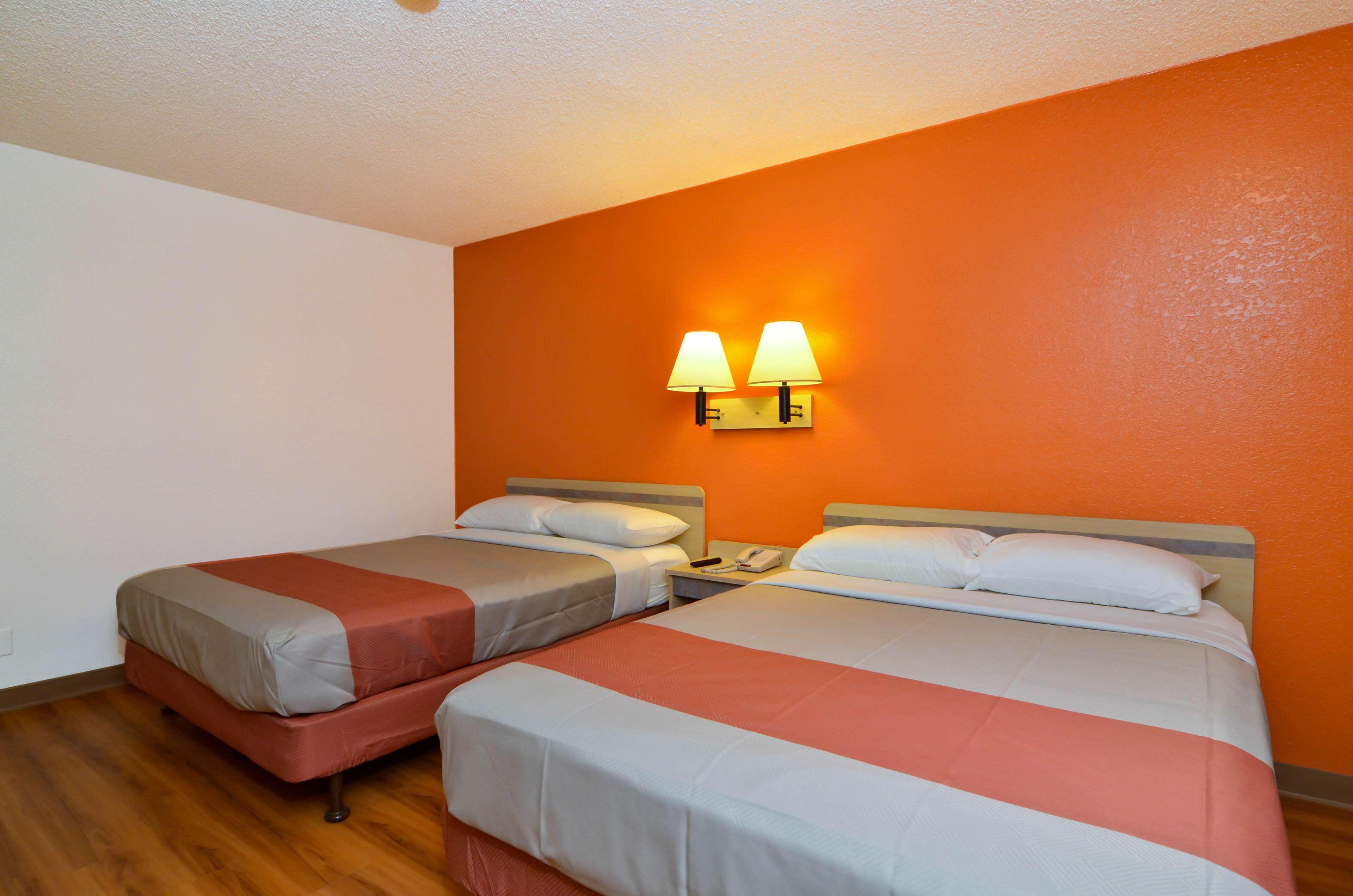 A hotel room with two beds, each with a pillow and a blanket. There are two wall-mounted lamps on the orange wall.