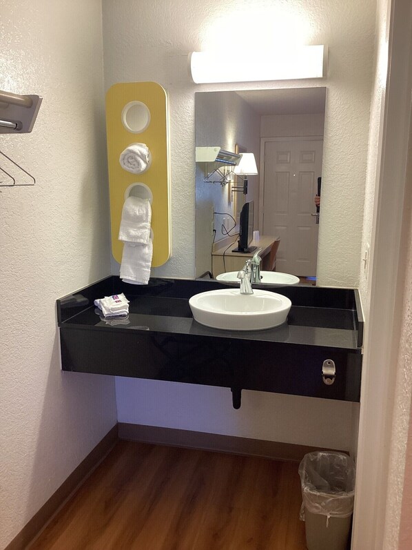 A bathroom with a black countertop, white sink, and a mirror. There is a towel rack with a yellow accent, a trash can, and a door in the background.