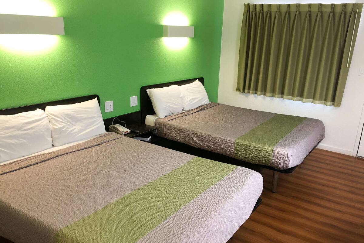Habitación de hotel con dos camas, paredes verdes, suelo de madera y una ventana con cortinas verdes.