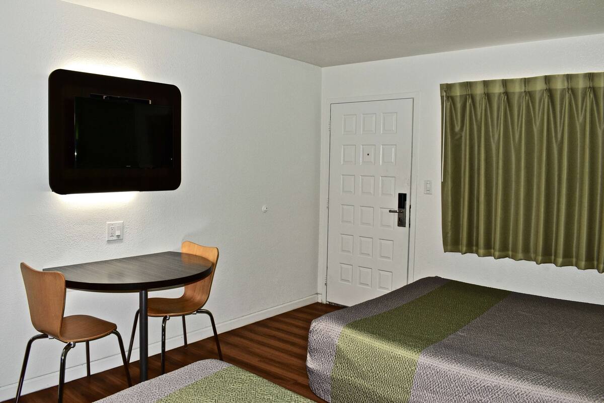 Una habitación con una cama individual, una mesita con dos sillas, un televisor montado en la pared y una puerta con un ventanal.