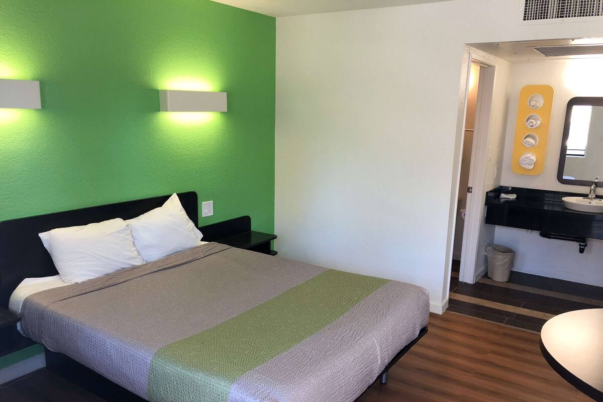 Habitación de hotel con cama, mesita de noche, pared verde, baño con lavabo y espejo, y un cubo de basura.