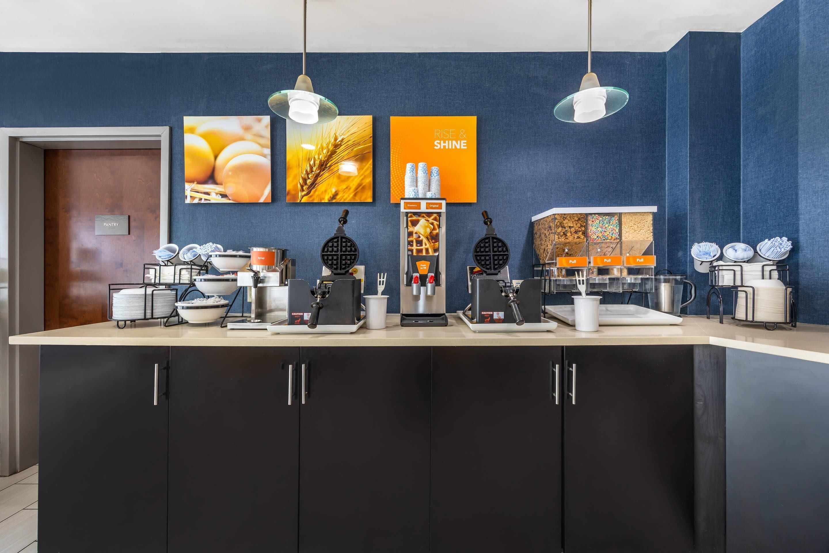 A kitchen area with waffle makers, coffee machines, and a sign that says "RISE & SHINE".
