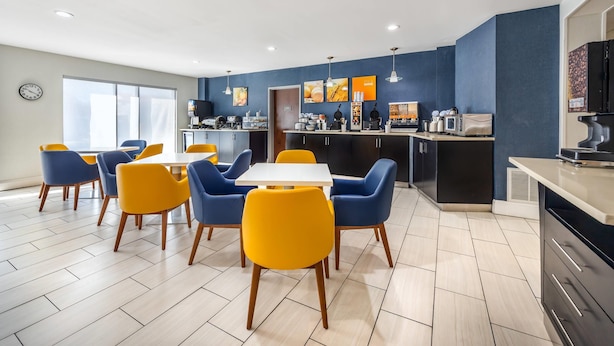 A modern dining area with a white table and yellow and blue chairs, a kitchen with appliances, and a clock on the wall.