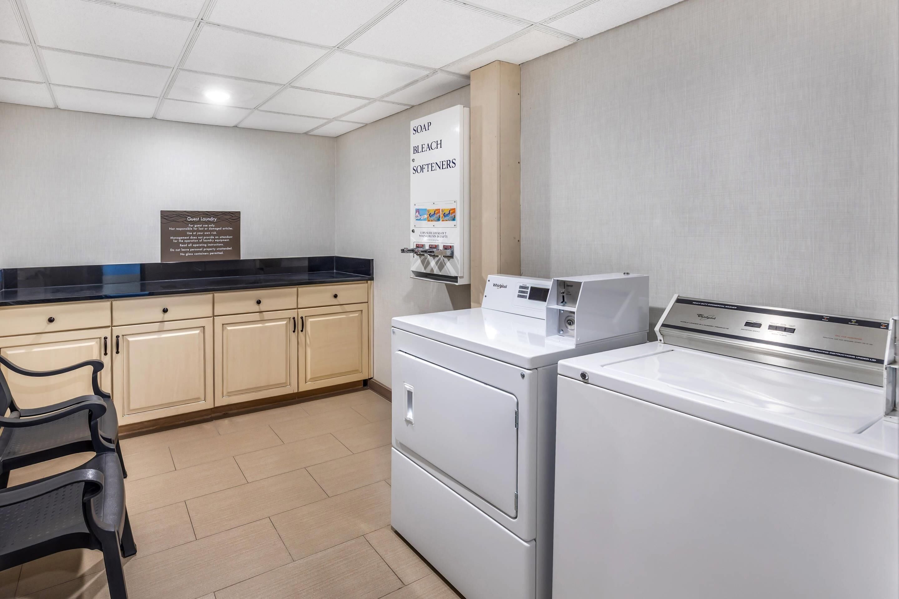 Een wasruimte met een wasbak, zeep, bleekmiddel en wasverzachter, een gastenwasberichtenbord, en een Whirlpool wasmachine en droger.