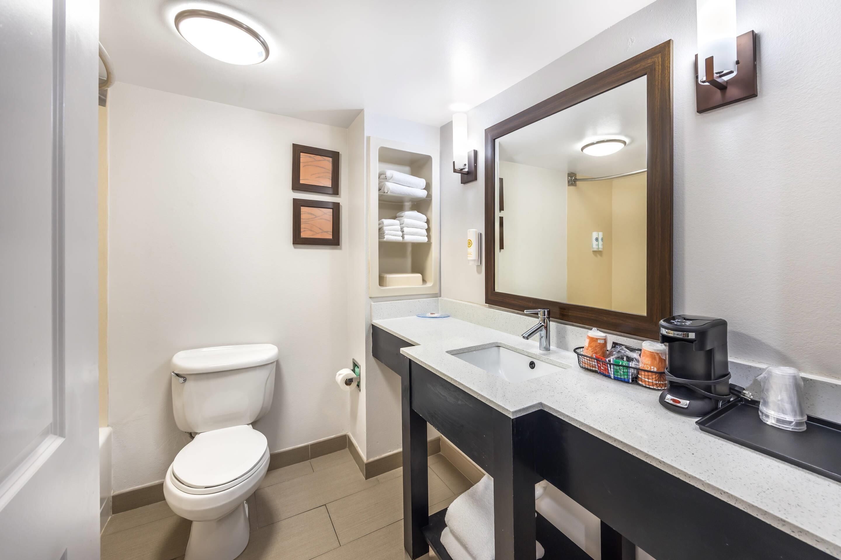 A modern bathroom with a white toilet, a large mirror, a sink with a black countertop, and a coffee maker.