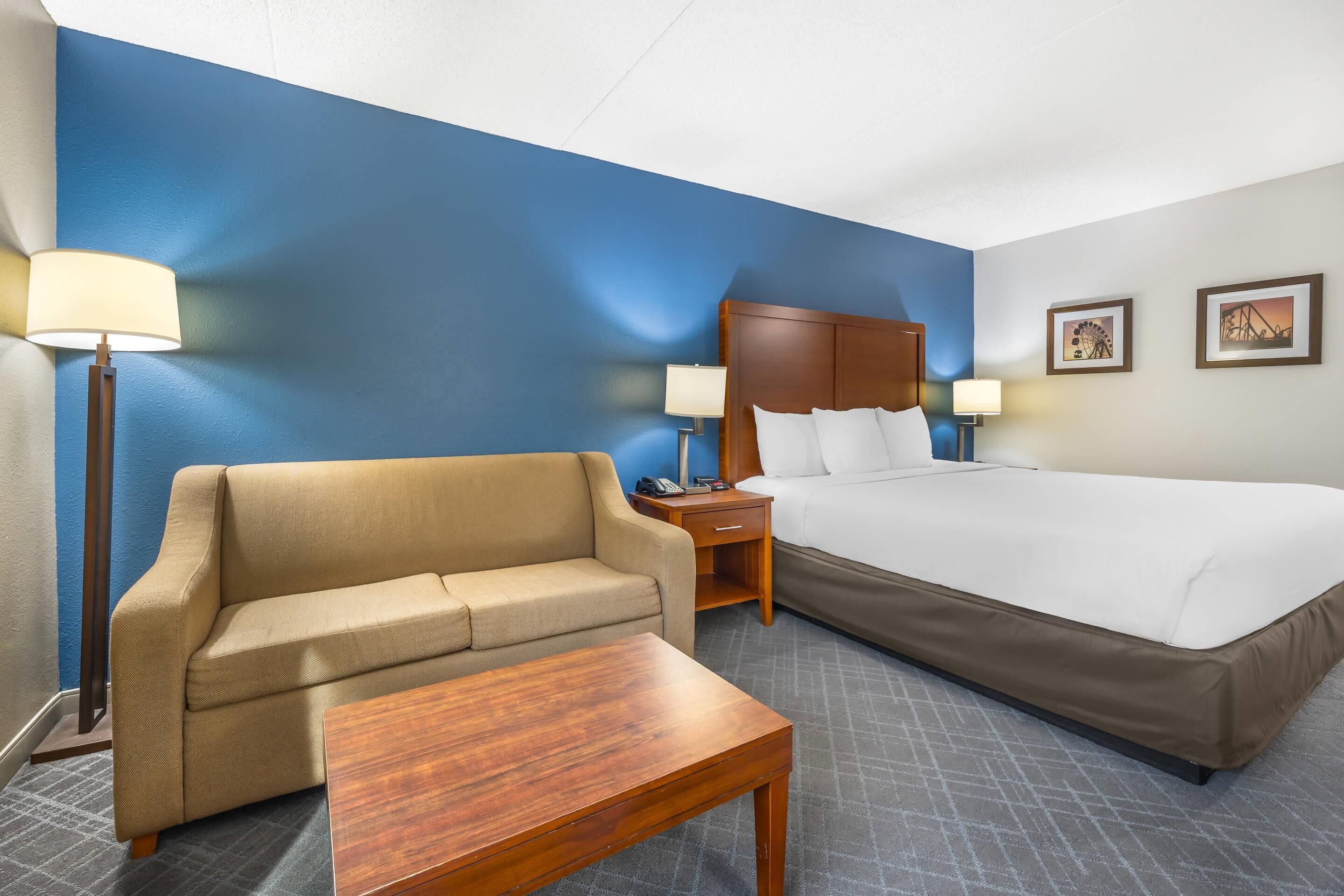 Habitación de hotel con cama, sofá, una mesa de madera, mesita de noche con lámpara y dos lámparas de pared.