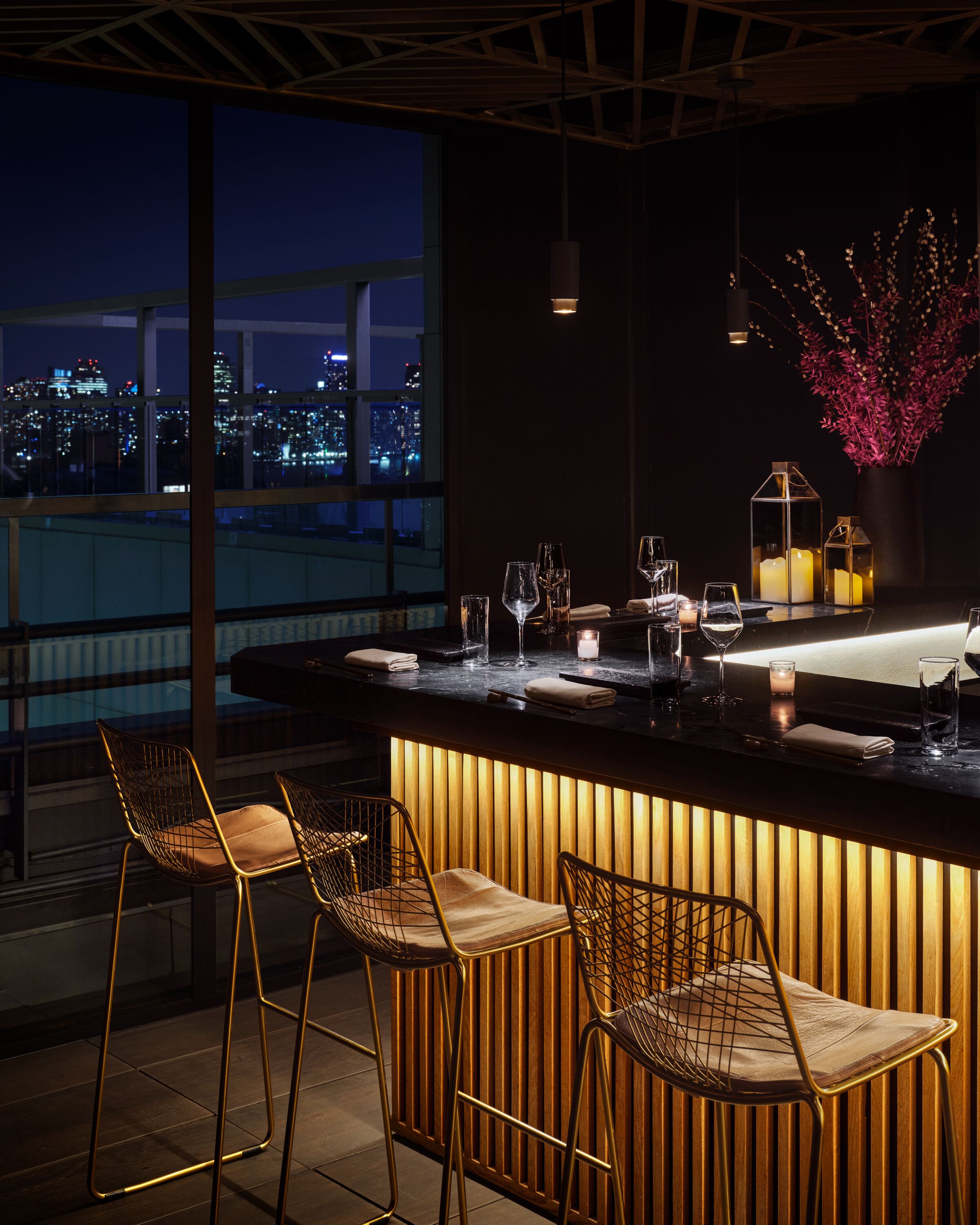 Un bar moderne offrant une vue sur la ville, avec un comptoir en bois, trois tabourets de bar en fil de fer et des bougies décoratives.